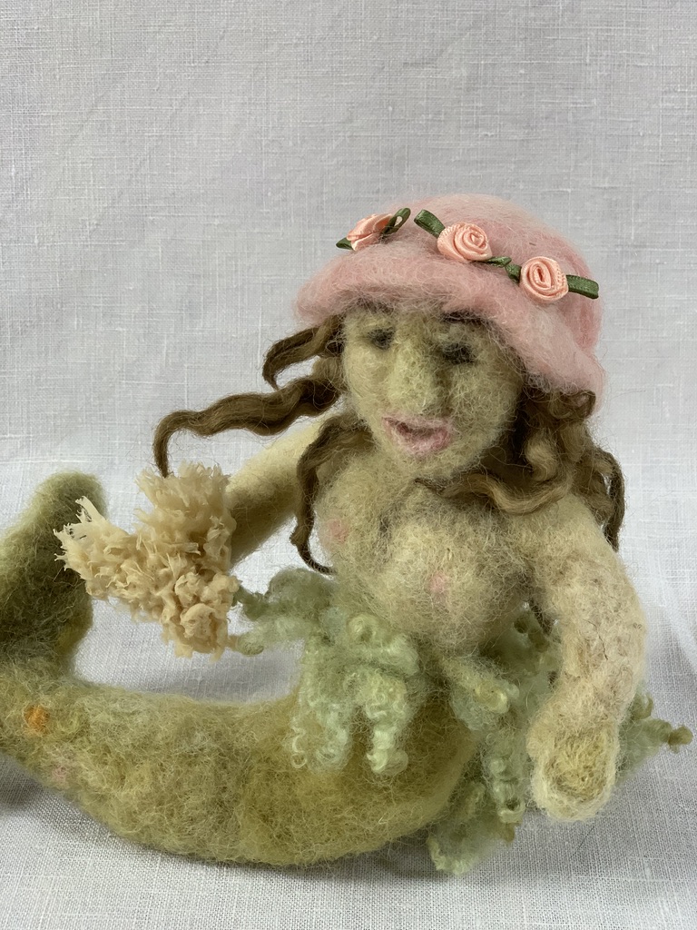Needle-felted mermaid wearing her new pink flowered hat, holding a sponge.